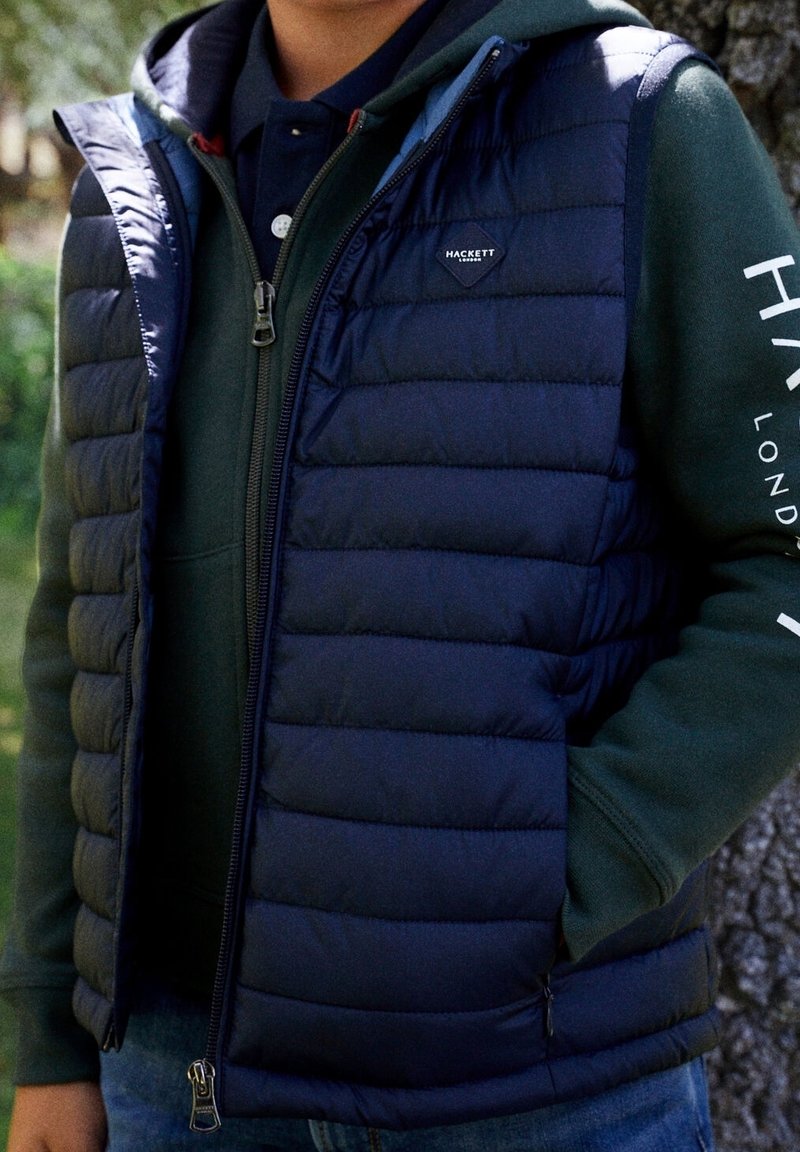 Granatowa pikowana kamizelka z zamkiem błyskawicznym i bocznymi kieszeniami, noszona na zielonej bluzie z kapturem. Na lewej piersi znajduje się logo, a materiał ma gładką teksturę.