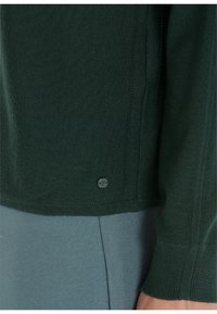 Maglione lavorato a maglia verde scuro con polsini e orlo a coste. Presenta un piccolo bottone con logo sulla cucitura inferiore. Tessuto leggero e dalle texture. 
