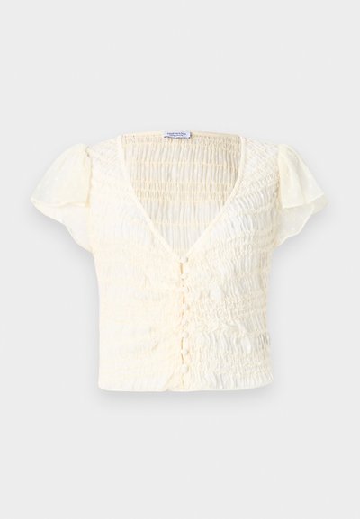Top curto de cor creme com textura franzida, decote em V e fecho de botão. Tecido leve com mangas ligeiramente volumosas.