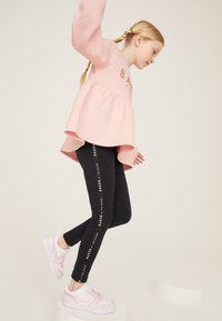 Rozā džemperis ar izvērstu maliņu un zelta uzrakstu, kombinēts ar melnām legingām ar baltu zīmola tekstu, papildināts ar rozā un baltām sporta kurpēm.