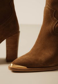 Ghete maro din piele întoarsă cu tocuri din lemn suprapuse și capă metalică aurie pe un fundal neutru.