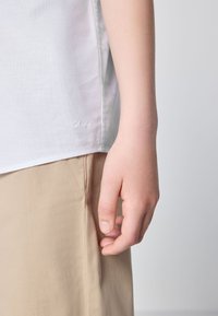 Close-up of a hand resting beside beige pants and a white shirt with a small embroidered logo near the hem.