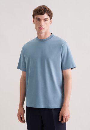 Junger Mann mit hellbraunen Haaren, trägt ein schlichtes hellblaues kurzärmeliges T-Shirt und dunkle Hosen, steht vor einem neutralen Hintergrund.