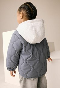 Veste grise matelassée avec capuche gris clair, arborant un motif ondulé et des poignets élastiques, portée sur un pantalon en denim ample.