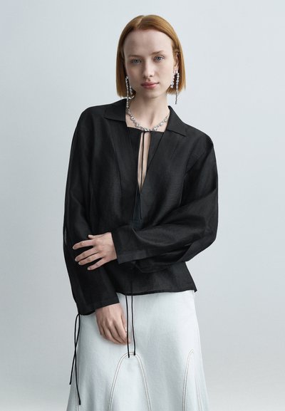 Blouse noire légère avec col et fermeture à nouer, dotée de manches longues. Portée avec une jupe blanche agrémentée de détails cousus.
