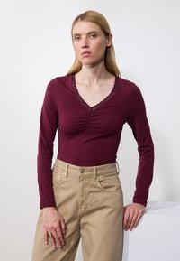 Burgundy long-sleeve top with a V-neck and lace trim, paired with light beige high-waisted pants, smooth texture, simple design.