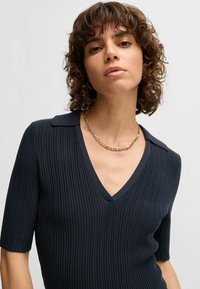 Ciemnoniebieski, prążkowany top z dekoltem w serek i krótkimi rękawami. Posiada klasyczny kołnierzyk i delikatny połysk. Złoty naszyjnik podkreśla linię dekoltu.