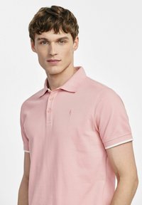 Jasnoróżowa koszulka polo z fakturowanej tkaniny, z trzema guzikami, kontrastującymi białymi akcentami na rękawach. Małe logo na klatce piersiowej.