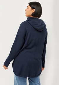 Marineblauwe hooded sweatshirt met een lange, relaxte pasvorm, voorzien van een zachte textuur en zijopeningen, gedragen over lichtblauwe jeans.