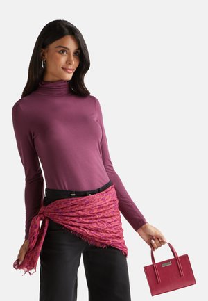 Lila Langarmoberteil, rosa gemusterter Schal um die Taille gebunden, schwarze Hose, hält eine kleine rote Handtasche mit Griffen. Strukturierte Materialien sichtbar.