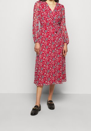 Kvinde iført en rød blomsterwrap-kjole med lange ærmer og sorte loafers med guldkæder, stående mod en ensfarvet lys baggrund.
