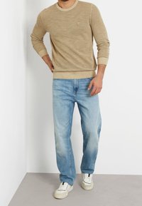 Uomo che indossa un maglione beige a trama, jeans blu chiaro a gamba dritta e sneakers bianche, in piedi contro un muro bianco.