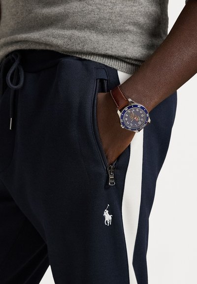 Jūras spēku sporta bikses ar baltām sānu līnijām un rāvējslēdzēja kabatu. Uz rokas ir sudraba pulkstenis ar zilu ciparnīcu un brūnu ādas siksniņu.