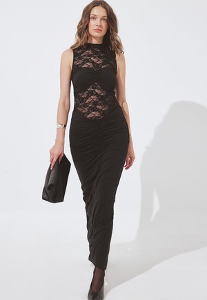 Kobieta w czarnej koronkowej sukience bez rękawów z marszczeniami, idąca, trzymająca czarną kopertówkę, ubrana w czarne szpilki i złotą bransoletkę.