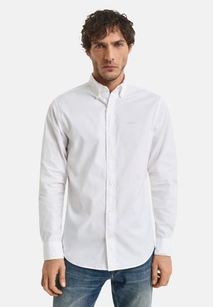 Weißes Button-Up-Hemd aus Baumwolle, mit klassischem Kragen, langen Ärmeln und einem dezenten Logo auf der linken Seite. Getragen mit blauen Jeans.