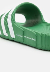Gröna Adidas flip-flops med vita ränder och texturerad sula, med texten "MÄRKET MED TRE RÄNDER" på sidan.