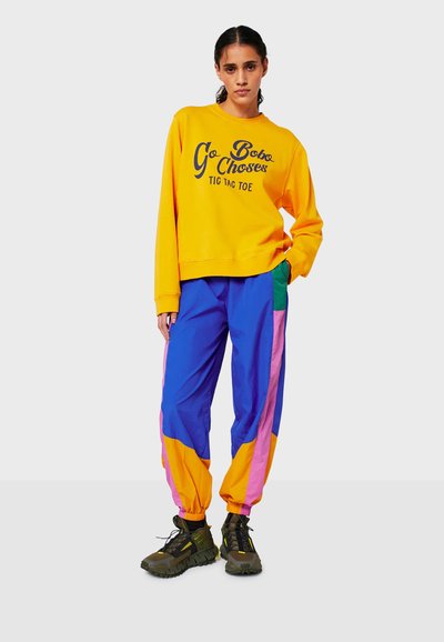 Felpa gialla brillante con testo blu navy, abbinata a pantaloni baggy multicolore blu, rosa, arancione e verde. Scarpe da trekking nere con dettagli gialli.