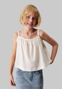 Weißes ärmelloses Top mit geripptem Textur und gerafftem Ausschnitt, mit einem kleinen grauen Logo auf der Brust, kombiniert mit hellblauer Jeans.