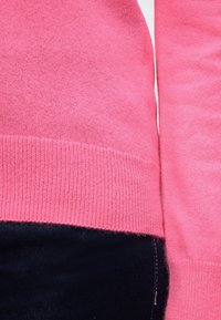 Gros plan d'une personne portant un pull en tricot rose vif et un pantalon sombre, montrant la texture et les détails des coutures du vêtement.