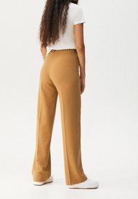 Spodnie o szerokich nogawkach i wysokim stanie w kolorze beżowym, wykonane z miękkiej tkaniny. Charakteryzują się gładką fakturą i subtelnym przeszyciem z tyłu. Noszone z białymi trampkami.