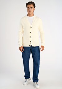 Krämfärgad ribbad kavaj med V-ringning, fem svarta knappar, bära över en vit T-shirt. Matchad med blå denimjeans och vita sneakers.