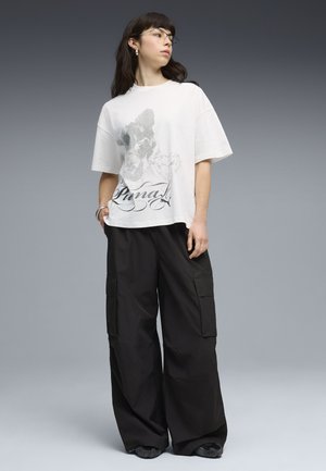 Frau mit dunklem Haar und Brille, trägt ein weißes kurzärmliges T-Shirt mit Grafik und lockere schwarze Cargo-Hosen vor einem grauen Hintergrund.