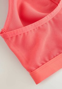 Detaliu al unui sutien sport roz coral, cu material textil, prezentând un panou din plasă cu cusături întărite și o bandă elastică pe un fundal deschis.