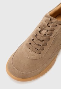 Sapatilhas em camurça castanho claro com bico arredondado, atacadores tonais, laterais texturizadas e sola de borracha. Apresenta uma marcação discreta na lateral.