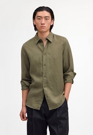 Ung mand med sort hår iført en olivengrøn skjorte med knapper og opkrogede ærmer samt sorte bukser, stående foran en ensfarvet baggrund.
