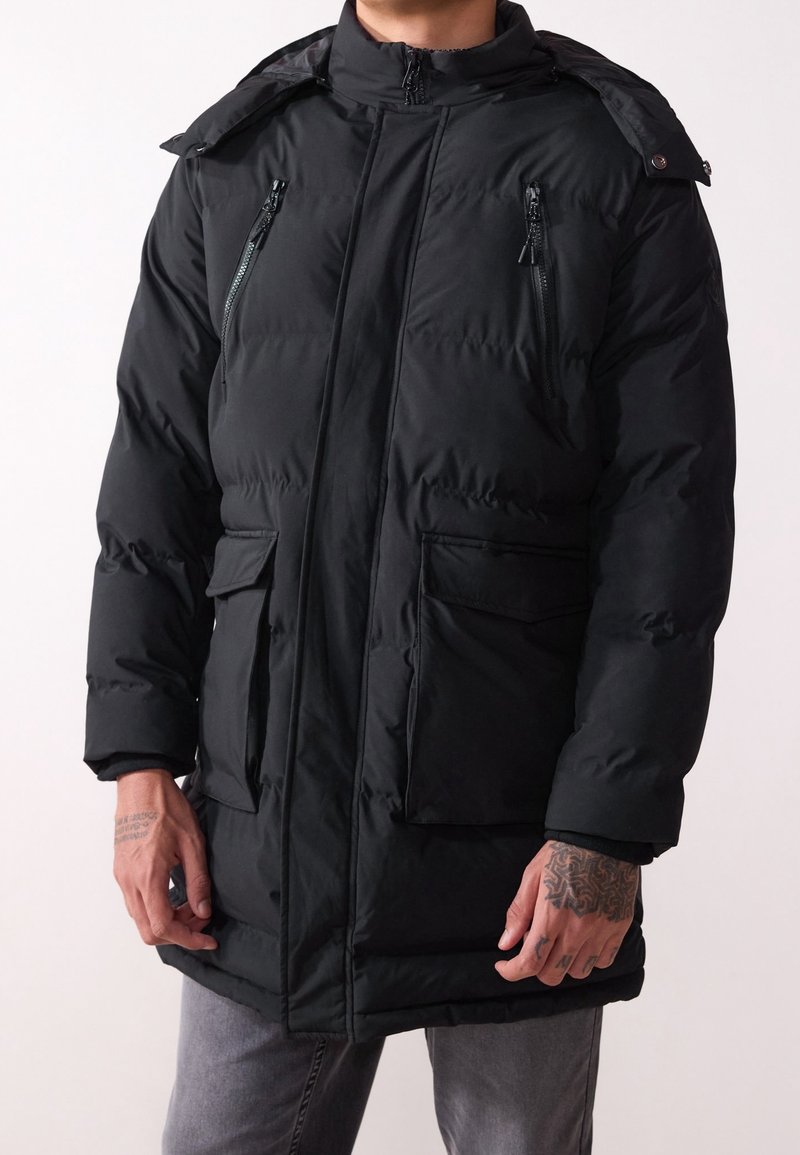Homme portant un manteau d'hiver noir matelassé avec des poches poitrine zippées, des poches à rabat à la taille et un col montant, debout devant un fond uni.