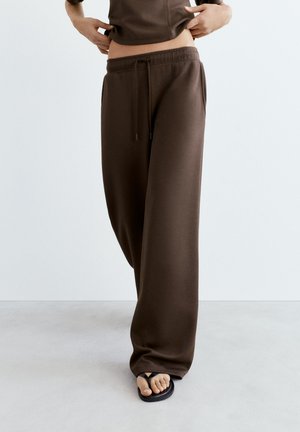 Persona che indossa pantaloni della tuta marroni larghi e infradito nere, sollevando leggermente una maglietta marrone per mostrare la zona della vita.