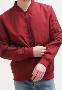 Rote Bomberjacke aus Nylon mit einem Reißverschluss vorne, gerippten Bündchen und einer mit Reißverschluss versehenen Tasche am linken Ärmel. Einfaches und funktionales Design.