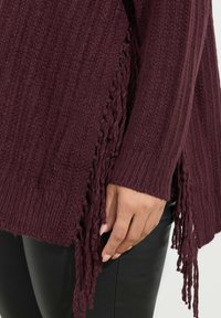 Bordowy sweter wykonany na drutach z teksturą ściągacza, z frędzlami na dolnym brzegu. Otaczający czarny materiał nie jest widoczny.