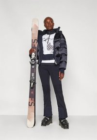 Síruházat: fekete pufi kabát, bordázott hatású sötétkék nadrág és fehér mintás pulóver; sífelszerelést tart a kezében.