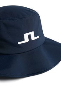 Marinblå bucket hat tillverkad av slätt, vattentätt tyg. Har en framträdande vit logotyp på framsidan. Mjuk textur och sydda kanter.