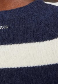 Pull à rayures bleu marine et crème, avec un col rond, un tricot texturé et un accent logo argent près du col.