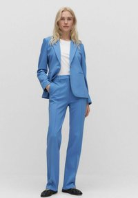 Ljusblå kostymset med en skräddarsydd kavaj och raka byxor, i kombination med en vit skjorta och svarta skor. Slät tyg, modern passform.