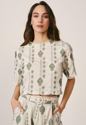 Mujer con cabello largo y castaño que lleva un conjunto de top y pantalones crema con patrones florales bordados en verde y un cinturón a juego, manos en los bolsillos.