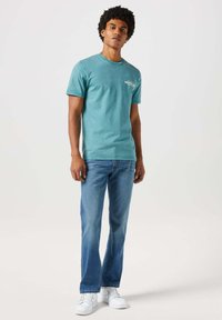T-shirt in cotone verde acqua con un piccolo logo bianco sul petto, abbinata a jeans in denim blu chiaro e scarpe da ginnastica bianche. Vestibilità casual.