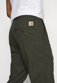 Olijfgroene broek van stevig materiaal, met een achterzak met knoopsluiting en een vierkant Carhartt logo patch.