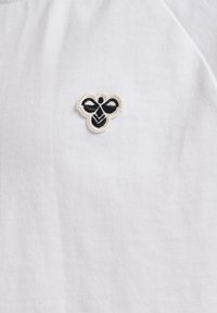 Chemise en tissu blanc avec un petit motif de abeille brodé en noir et crème sur la poitrine gauche, mettant en valeur des points de couture détaillés et une texture.