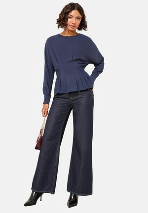Mörkblå peplumtop med långa ärmar, texturerat tyg och veck i midjan, kombinerad med vida mörk denimjeans och svarta klackar.