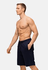 Muskelbepackter Mann, der seitlich steht und dunkelblaue Shorts mit Kordelzug trägt, mit einer Hand in der Tasche vor einem schlichten weißen Hintergrund.