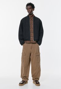 Crna bomber jakna preko smeđeg kariranog košulje, upaired s bež cargo hlačama koje imaju više džepova. Smeđe cipele na vezanje zaokružuju izgled.