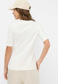 Top bianco a righe con maniche corte e scollatura rotonda, abbinato a pantaloni beige chiaro. Capelli mossi e indossa un cappello chiaro.