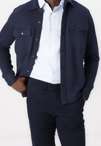 Mörkblå skjortjacka med knappar, med texturerat tyg och två framfickor, lager-på-lager över en ljusblå skjorta med krage och mörka byxor.