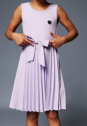 Enfant portant une robe lavande plissée sans manches avec un nœud à la taille et une petite épingle noire en forme de cœur sur la poitrine.