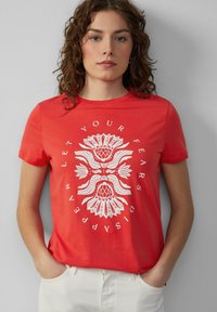 T-shirt en coton rouge à manches courtes ornée d'un motif floral blanc et d'un design de texte circulaire. À assortir avec un pantalon de couleur claire.