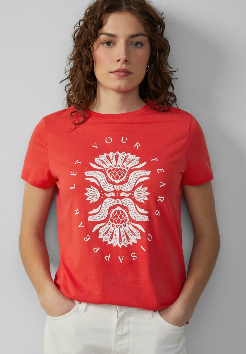 T-shirt en coton rouge à manches courtes ornée d'un motif floral blanc et d'un design de texte circulaire. À assortir avec un pantalon de couleur claire.