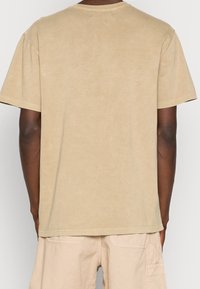 Osoba v jednoduchém béžovém tričku s krátkým rukávem a světle béžových cargo šortkách, zobrazená zezadu na neutrálním pozadí.